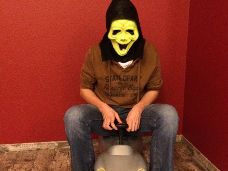 Maskierter Fahrer auf Spielzeugauto Mann mit gelber Gruselmaske und Kapuze sitzt auf einem grauen Spielzeugauto.